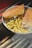 A metal tray holds a sandwich with a seeded crust, filled with layers of grilled meat and melted cheese, accompanied by a generous portion of seasoned French fries sprinkled with herbs. The background includes cane-like sticks placed on a dark surface.