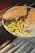 A metal tray holds a sandwich with a seeded crust, filled with layers of grilled meat and melted cheese, accompanied by a generous portion of seasoned French fries sprinkled with herbs. The background includes cane-like sticks placed on a dark surface.