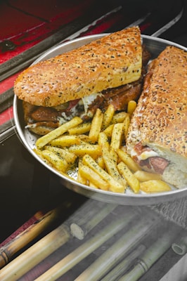 A metal tray holds a sandwich with a seeded crust, filled with layers of grilled meat and melted cheese, accompanied by a generous portion of seasoned French fries sprinkled with herbs. The background includes cane-like sticks placed on a dark surface.