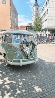 A vintage Volkswagen van, decorated with a heart-shaped floral arrangement on the front grille, is parked in a cobblestone plaza. The setting includes modern and historic buildings, with a clock tower in the background. Nearby, there are several bicycles parked and a few pedestrians can be seen.