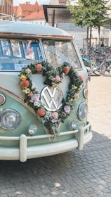 A vintage Volkswagen van decorated with a heart-shaped floral wreath on its front. The wreath is composed of various colorful flowers, including pink and orange blooms. The van is parked on a cobblestone street with bicycles visible in the background, conveying a quaint and charming street scene.