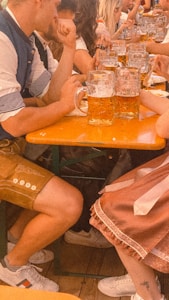 A group of people are seated around a long wooden table, enjoying large mugs of beer. Many are wearing traditional Bavarian attire, such as lederhosen and dirndl dresses. The atmosphere appears festive and lively.