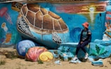 A colorful mural of a sea turtle surrounded by ocean-themed elements such as jellyfish, seaweed, and shells. A person wearing a red hat and casual clothing is leaning against the painted wall, adding a human element to the vibrant scene.