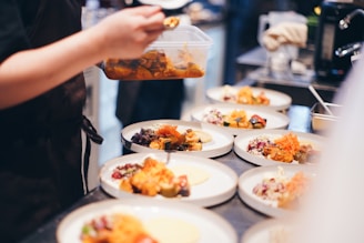 A person is plating food in a kitchen, preparing several dishes with vibrant ingredients. Various plates are arranged on a countertop, each containing a variety of colorful foods, such as vegetables and garnishes.