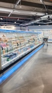 A supermarket aisle contains a long refrigerated display filled with various packaged products. Bright lighting illuminates the area, highlighting items such as meats and dairy. The floor is polished concrete, and a shopping cart is visible in the distance.