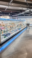 A supermarket aisle contains a long refrigerated display filled with various packaged products. Bright lighting illuminates the area, highlighting items such as meats and dairy. The floor is polished concrete, and a shopping cart is visible in the distance.
