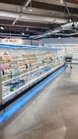 A supermarket aisle contains a long refrigerated display filled with various packaged products. Bright lighting illuminates the area, highlighting items such as meats and dairy. The floor is polished concrete, and a shopping cart is visible in the distance.