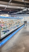 A supermarket aisle contains a long refrigerated display filled with various packaged products. Bright lighting illuminates the area, highlighting items such as meats and dairy. The floor is polished concrete, and a shopping cart is visible in the distance.
