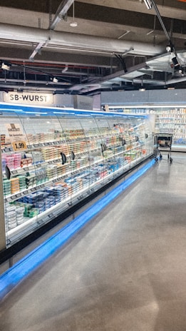 A supermarket aisle contains a long refrigerated display filled with various packaged products. Bright lighting illuminates the area, highlighting items such as meats and dairy. The floor is polished concrete, and a shopping cart is visible in the distance.