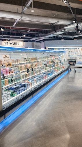 A supermarket aisle contains a long refrigerated display filled with various packaged products. Bright lighting illuminates the area, highlighting items such as meats and dairy. The floor is polished concrete, and a shopping cart is visible in the distance.