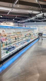 A supermarket aisle contains a long refrigerated display filled with various packaged products. Bright lighting illuminates the area, highlighting items such as meats and dairy. The floor is polished concrete, and a shopping cart is visible in the distance.