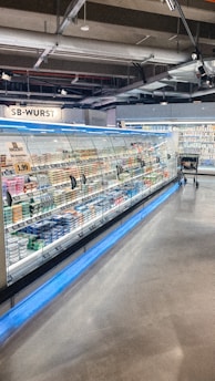 A supermarket aisle contains a long refrigerated display filled with various packaged products. Bright lighting illuminates the area, highlighting items such as meats and dairy. The floor is polished concrete, and a shopping cart is visible in the distance.