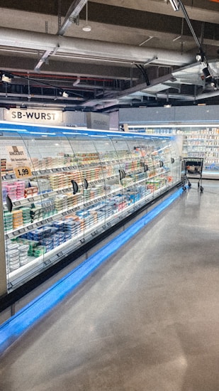 A supermarket aisle contains a long refrigerated display filled with various packaged products. Bright lighting illuminates the area, highlighting items such as meats and dairy. The floor is polished concrete, and a shopping cart is visible in the distance.