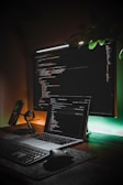 A workspace featuring a large monitor displaying code is complemented by a laptop also showing a code editor. The setup includes a mechanical keyboard, a wireless mouse, a smartphone on a stand, and ambient lighting creating a warm glow. A leafy plant adds a touch of greenery.