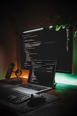 A workspace featuring a large monitor displaying code is complemented by a laptop also showing a code editor. The setup includes a mechanical keyboard, a wireless mouse, a smartphone on a stand, and ambient lighting creating a warm glow. A leafy plant adds a touch of greenery.