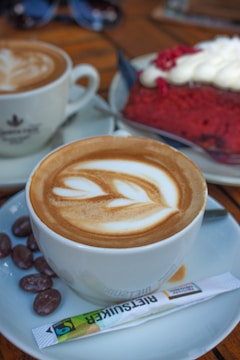 A latte with intricate latte art sits on a white saucer, accompanied by chocolate-covered nuts and a sugar packet labeled 'RIESZUCKER.' In the background, a piece of red velvet cake topped with cream is visible next to another coffee cup.
