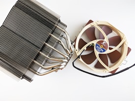A large CPU cooler featuring a dark metal heat sink with numerous fins and several heat pipes. Attached to it is a tan-colored cooling fan with brown blades, connected by a black wire. The fan's hub has a circular label with technical details.