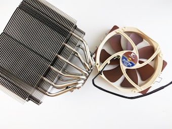 A large CPU cooler featuring a dark metal heat sink with numerous fins and several heat pipes. Attached to it is a tan-colored cooling fan with brown blades, connected by a black wire. The fan's hub has a circular label with technical details.