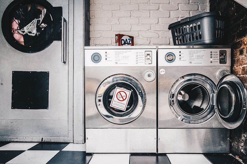 A laundromat setting featuring two industrial-style washing machines with metal finishes. One machine door is open, revealing the drum inside, while the other has a 'No Smoking' sign attached to it. There's a detergent box labeled 'Daz' placed on top of the machines beside a plastic laundry basket. The floor has a black and white checkered pattern, and a brick wall is visible in the background.