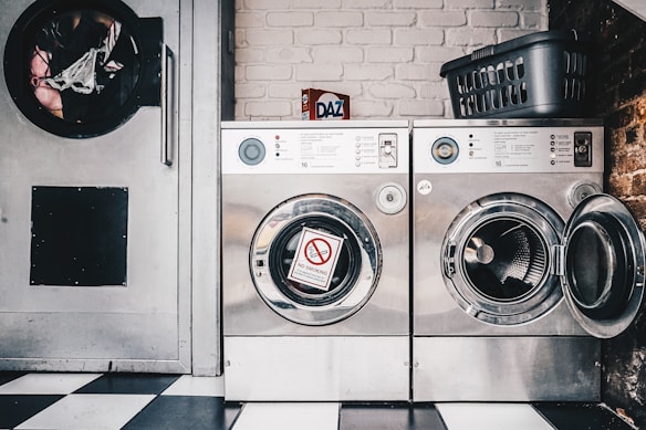 A laundromat setting featuring two industrial-style washing machines with metal finishes. One machine door is open, revealing the drum inside, while the other has a 'No Smoking' sign attached to it. There's a detergent box labeled 'Daz' placed on top of the machines beside a plastic laundry basket. The floor has a black and white checkered pattern, and a brick wall is visible in the background.