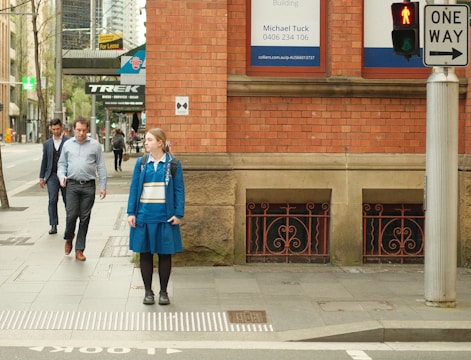 A young woman in a blue school uniform stands on a city sidewalk near a brick building, waiting at a crosswalk with a red pedestrian signal. Two men in business attire walk towards her from the opposite direction. Signs for a bicycle shop and a 'For Lease' ad are visible in the background.