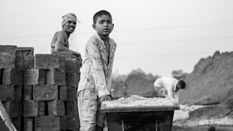 A young boy in work attire is pushing a wheelbarrow filled with sand or dirt. Behind him stand stacks of bricks, and to the left, a man leans on the stack, observing. In the background, another person is bent over, seemingly engaged in work, with mounds of earth surrounding the scene.