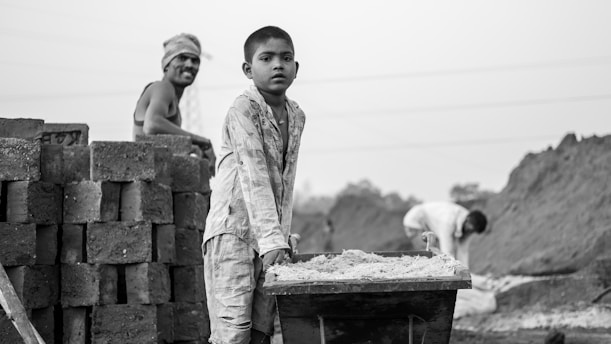 A young boy in work attire is pushing a wheelbarrow filled with sand or dirt. Behind him stand stacks of bricks, and to the left, a man leans on the stack, observing. In the background, another person is bent over, seemingly engaged in work, with mounds of earth surrounding the scene.
