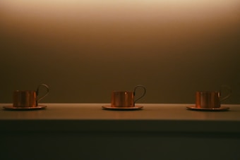 Three copper mugs with handles are aligned on top of a surface, all placed on matching copper saucers. The scene is set against a softly lit background with a warm, muted palette.