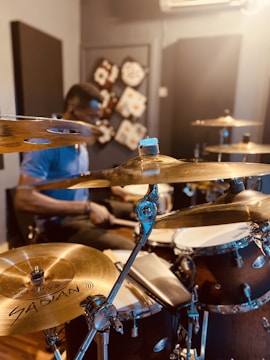 A musician is playing a drum set in a music studio. The focus is on the cymbals and other drum components, with soft lighting creating a concentrated and dynamic atmosphere. Behind the drummer, there are acoustic panels with abstract designs on the walls.