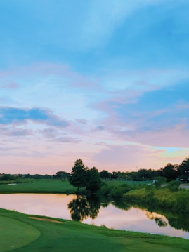 A serene landscape with a reflective body of water surrounded by lush greenery and trees. The sky is expansive with soft pastel hues of pink and blue, suggesting a sunset or sunrise. The water mirrors the trees and sky, creating a peaceful, symmetrical scene.