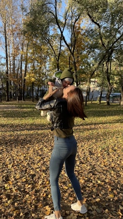 A smiling mother holding her baby in a sunlit park, surrounded by autumn leaves