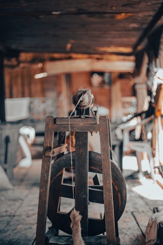The image depicts an old wooden spinning wheel placed centrally in an indoor setting. The background is slightly blurred but hints at a rustic, perhaps workshop or barn-like environment with various tools and objects scattered around.