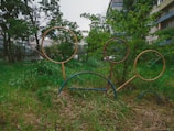 Outdoor playground rings on a grassy area surrounded by trees and apartment buildings. The rings are metal and stand at different heights, with greenery filling much of the scene.