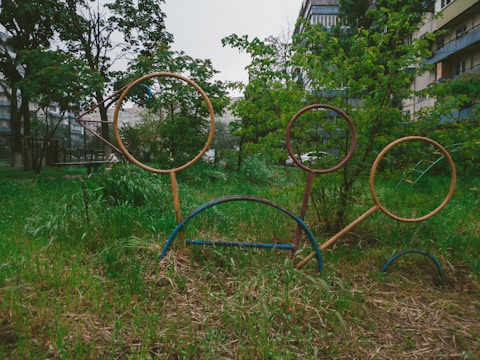 Outdoor playground rings on a grassy area surrounded by trees and apartment buildings. The rings are metal and stand at different heights, with greenery filling much of the scene.
