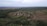 Aerial view of a rural landscape featuring a small village surrounded by dense forests and fields. The village consists of scattered houses and a network of dirt roads. The horizon is visible, with fields extending into the distance.