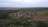 Aerial view of a rural landscape featuring a small village surrounded by dense forests and fields. The village consists of scattered houses and a network of dirt roads. The horizon is visible, with fields extending into the distance.