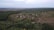 Aerial view of a rural landscape featuring a small village surrounded by dense forests and fields. The village consists of scattered houses and a network of dirt roads. The horizon is visible, with fields extending into the distance.