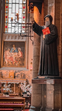 A statue of a hooded figure holding a red book stands beside ornate stone pillars. The background features a detailed religious painting depicting a biblical scene, situated beneath a colorful stained glass window. Below, a small altar is adorned with white flowers and candles.