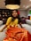 A young girl is sitting on a large grey sofa in a warmly lit room. She is wearing a traditional outfit with a yellow top and an orange skirt featuring golden patterns. A large red flower adorns her hair, and she is accessorized with golden bracelets.