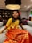 A young girl is sitting on a large grey sofa in a warmly lit room. She is wearing a traditional outfit with a yellow top and an orange skirt featuring golden patterns. A large red flower adorns her hair, and she is accessorized with golden bracelets.