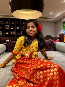 A young girl is sitting on a large grey sofa in a warmly lit room. She is wearing a traditional outfit with a yellow top and an orange skirt featuring golden patterns. A large red flower adorns her hair, and she is accessorized with golden bracelets.