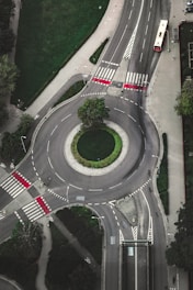 An aerial view of a roundabout with well-marked pedestrian crossings and distinct road lanes. Surrounding the roundabout are patches of green grass and trees. A red bus is visible on one of the roads exiting the roundabout, and bike lanes are marked alongside the roadways.