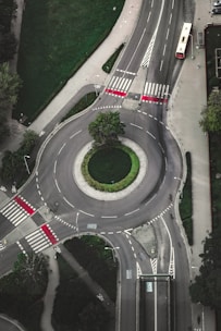 An aerial view of a roundabout with well-marked pedestrian crossings and distinct road lanes. Surrounding the roundabout are patches of green grass and trees. A red bus is visible on one of the roads exiting the roundabout, and bike lanes are marked alongside the roadways.
