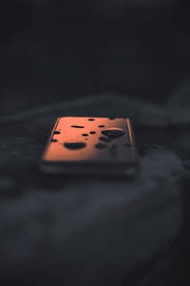 A smartphone lies on a dark surface with water droplets scattered across its screen. The screen reflects a warm orange glow, contrasting with the darker surroundings, creating a mysterious and moody atmosphere.
