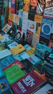 A variety of books are arranged in a cluttered, overlapping manner, showcasing a multitude of titles and colorful covers. The books are laid out horizontally and vertically, with genres ranging from self-help and fiction to non-fiction and essays.