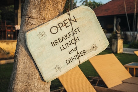 A weathered wooden sign is hanging on a tree, featuring the text 'OPEN BREAKFAST LUNCH and DINNER' along with floral designs. In the background, there are wicker lounge chairs, a grassy lawn, and a building with a reddish-brown roof.