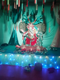 A detailed altar setup featuring a statue of Ganesha adorned with red garments and garlands. Positioned behind Ganesha is a large statue of another deity with multiple arms and large drum-shaped elements. The setting includes various offerings and decorative items like flowers, lamps, and cloths. Decorative string lights are draped along the front side.