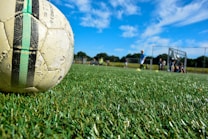 A well-used soccer ball lies on a lush green grass playing field under a bright blue sky with scattered clouds. In the background, a group of children in sportswear is visible, some standing and some sitting near a goalpost. The scene is situated outdoors, with a sense of motion and activity in a sports setting.
