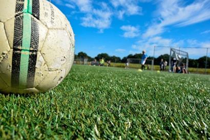 A well-used soccer ball lies on a lush green grass playing field under a bright blue sky with scattered clouds. In the background, a group of children in sportswear is visible, some standing and some sitting near a goalpost. The scene is situated outdoors, with a sense of motion and activity in a sports setting.