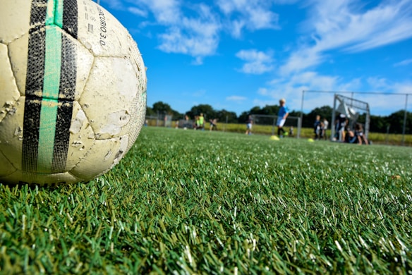 A well-used soccer ball lies on a lush green grass playing field under a bright blue sky with scattered clouds. In the background, a group of children in sportswear is visible, some standing and some sitting near a goalpost. The scene is situated outdoors, with a sense of motion and activity in a sports setting.